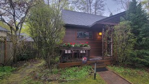 Une cabane en bois dotée d’un porche, entourée d’arbres et d’un jardin.