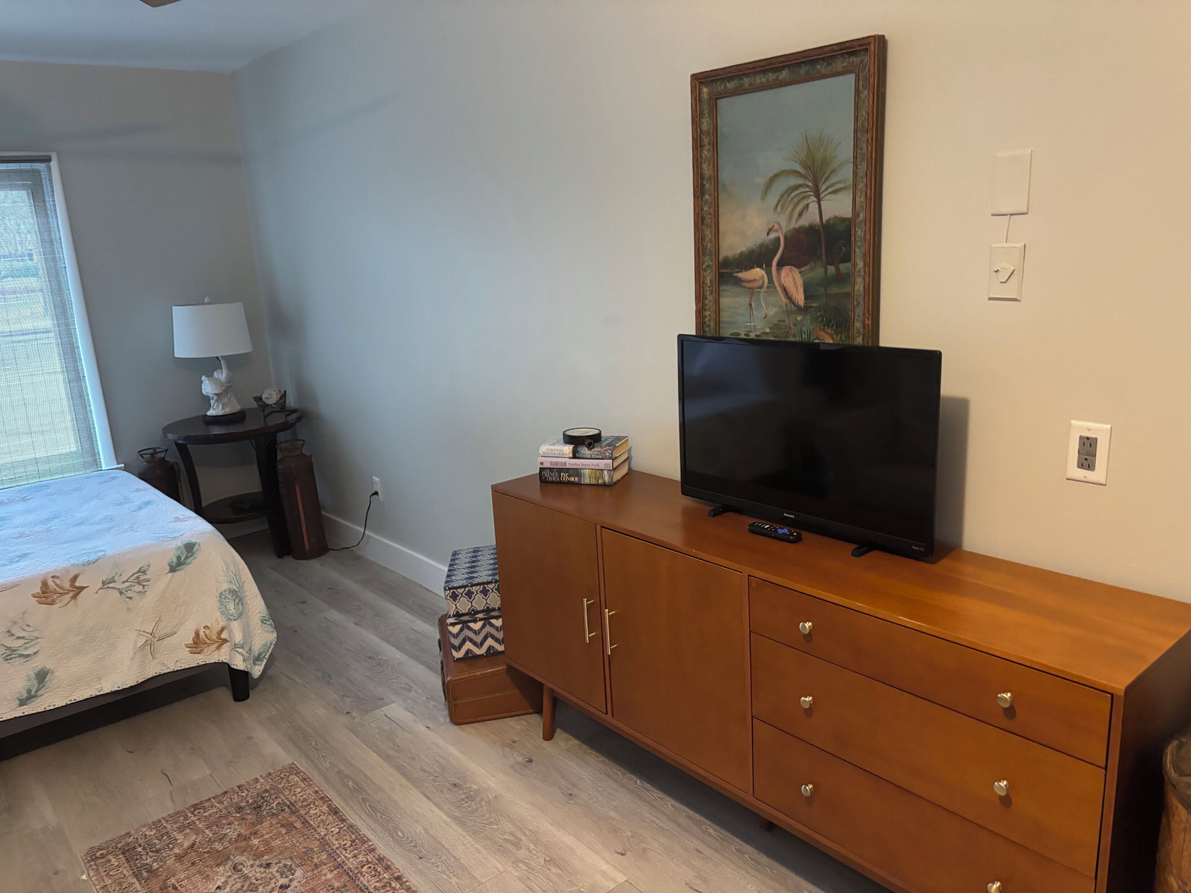 Een slaapkamer met een bed, een televisie op een houten dressoir, een lamp en een schilderij aan de muur.