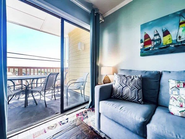 A living room with a sofa, a painting of sailboats, and a view through a glass door to an outdoor patio with chairs and a table.