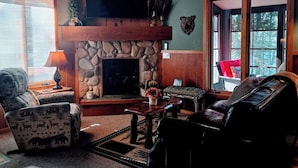 Een gezellige woonkamer met een stenen openhaard, een televisie erboven en een comfortabele zithoek met een bank en fauteuils.