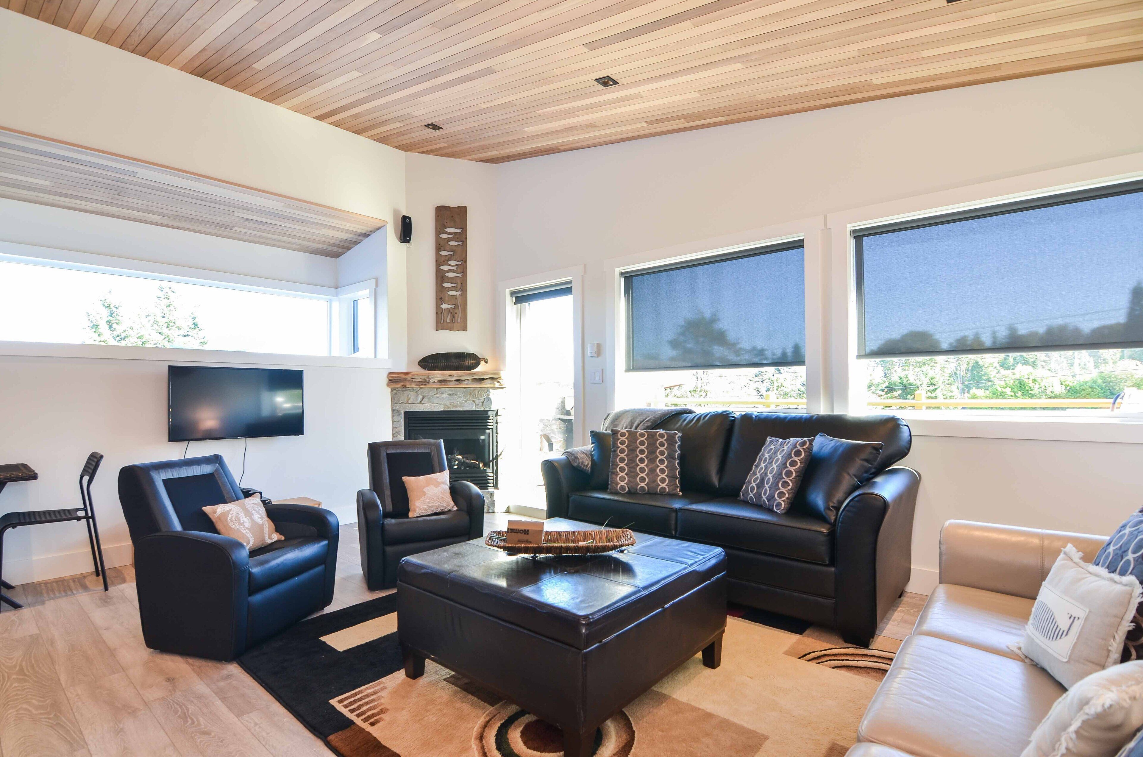 A modern living room with a fireplace, leather sofas, a flat-screen TV, and large windows.