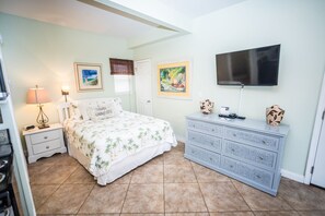 A bedroom with a bed, nightstand, dresser, TV, and artwork. - Crystal Beach Cottage, steps to beach & pool.  Close to shopping, restaurants! (Destin)