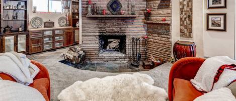 Una sala de estar acogedora con chimenea de piedra, sillones rojos y blancos, un alfombrero de pelo blanco y una estantería de madera con varios objetos decorativos.