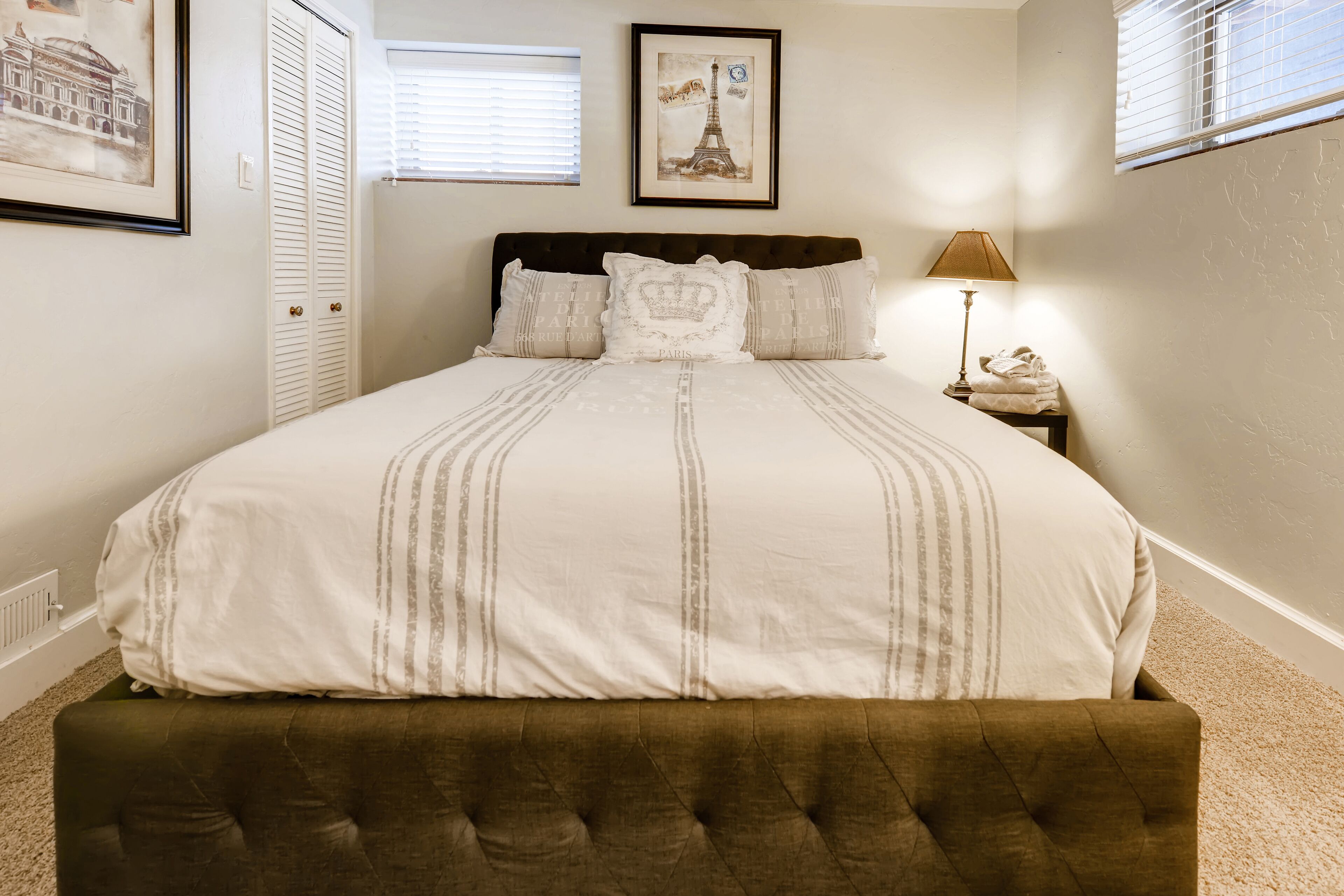 A bedroom with a large bed, a headboard, a nightstand with a lamp, and a window with blinds.