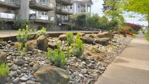 A multi-story residential building with balconies, a landscaped garden with rocks and plants, and a clear sky.