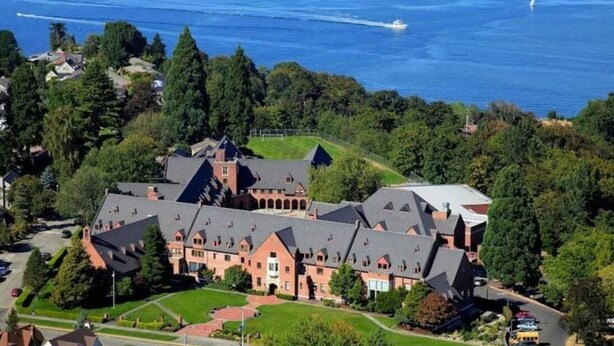 An aerial view of a large, historic building with multiple wings and a central courtyard, surrounded by lush greenery and a body of water.