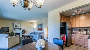 A modern living room with a fireplace, a dining table with a vase of flowers, and a kitchen with wooden cabinets and stainless steel appliances.