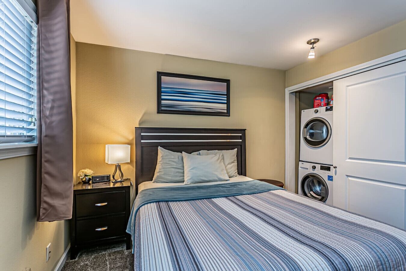 Habitación de hotel con cama, mesita de noche, lámpara, un cuadro enmarcado y lavadora secadora incorporada.