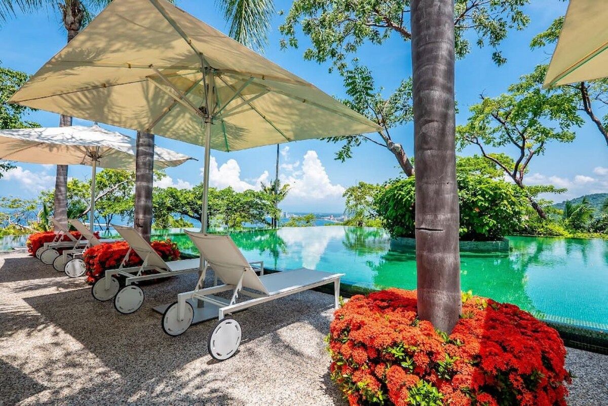 Un espace aménagé au bord de la piscine, avec des chaises longues et des parasols, entouré de verdure et offrant une vue dégagée sur l’eau.
