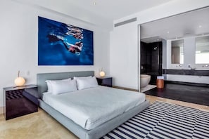 A modern bedroom with a large bed, a nightstand with a lamp, a wall-mounted artwork, and a bathroom with a large mirror.