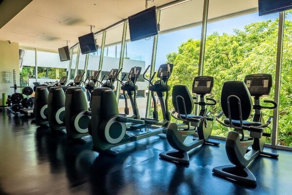 A gym with various exercise equipment, large windows, and a view of greenery outside.