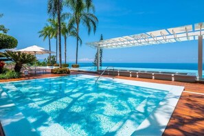 A pool with a view of the ocean, surrounded by palm trees and a covered seating area.