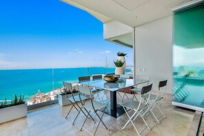 A balcony with a glass table and chairs, overlooking a coastal view.