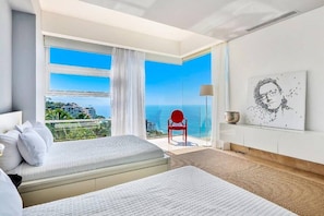 A hotel room with a bed, a red chair, a view of the ocean, and a painting on the wall.