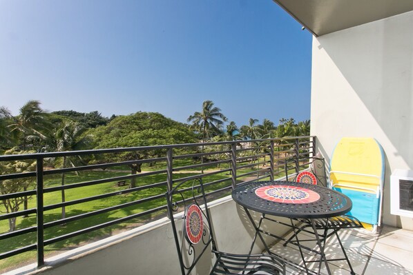 A balcony with a table and chairs, a view of a green park, and a palm tree.