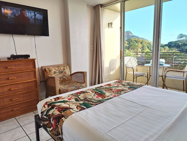 A hotel room with a bed, a dresser, a television, and a balcony with chairs and a table. - Ocean and Diamondhead view Queen Bed suite w/balcony - WAIKIKI GRAND HOTEL® 408 (Honolulu)
