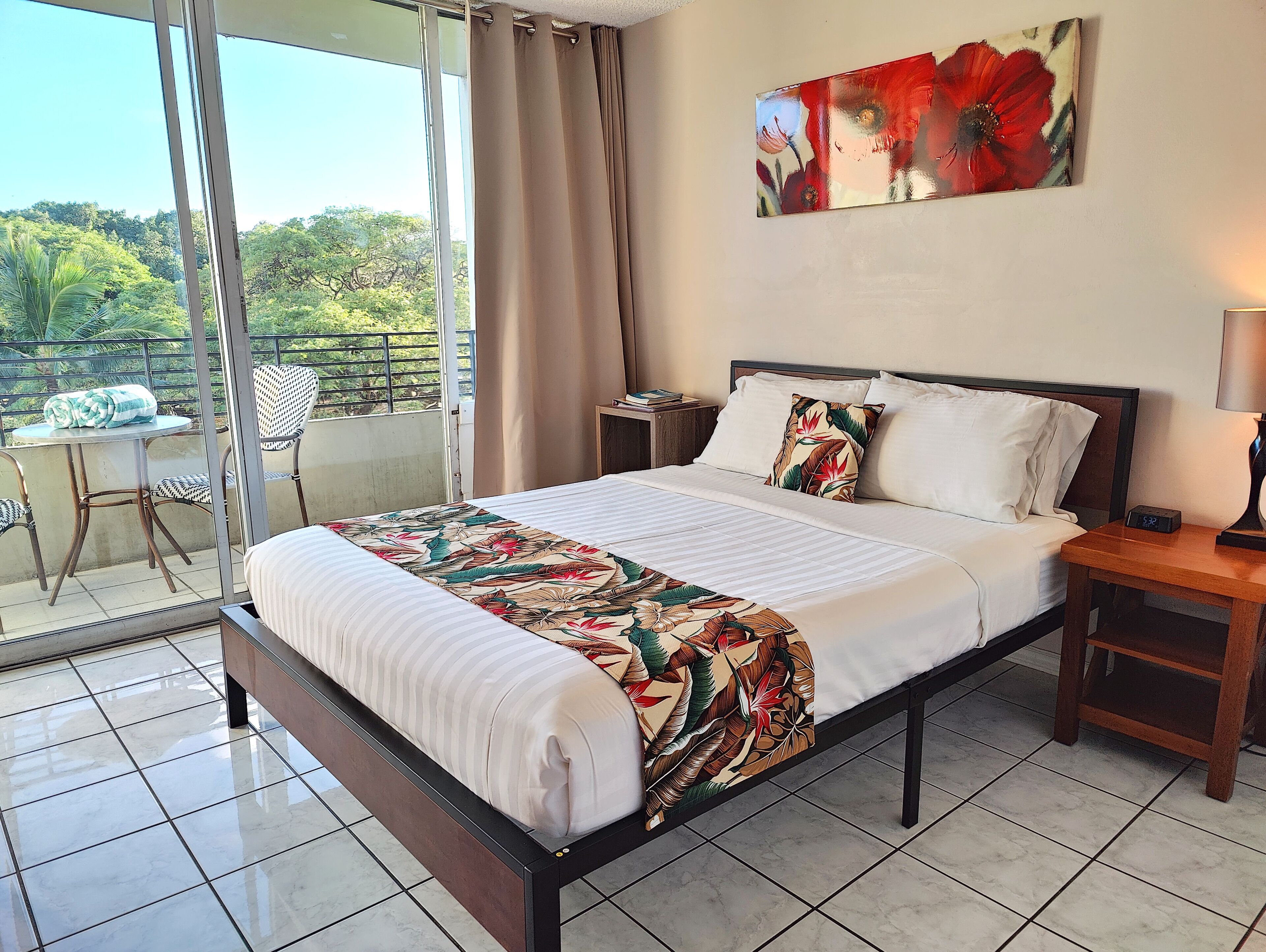 A hotel room with a bed, a nightstand, a table with chairs, and a view of greenery outside.