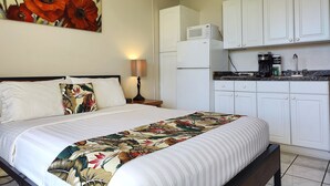 Un dormitorio con cama, mesita de noche, lámpara, un cuadro, y una cocina con armarios, refrigerador y cafetera.