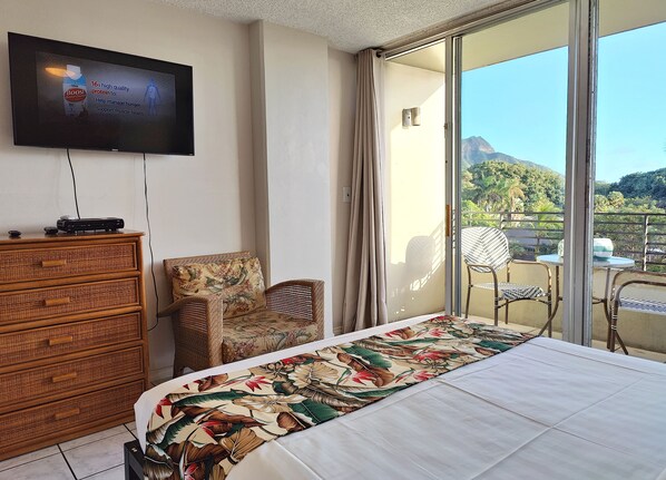 Habitación de hotel con cama, televisor, cómoda, una silla y balcón con vista a las montañas.