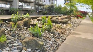 Un immeuble résidentiel à plusieurs étages doté de balcons, d’un jardin aménagé avec des rochers et des plantes, et d’un ciel dégagé.