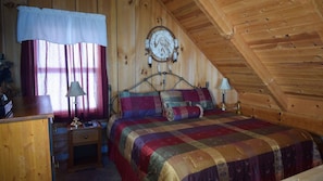 Ein gemütliches Zimmer mit Holzbalkendecke, einem Bett mit farbenfroher Steppdecke, einem Nachttisch mit Lampen und einem Fenster mit Vorhängen.