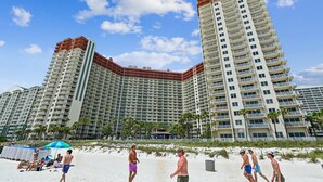 Un hotel de gran altura con múltiples balcones, una playa arenosa al frente y palmeras.