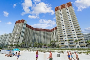 A high-rise hotel with multiple balconies, a sandy beach in the foreground, and palm trees.