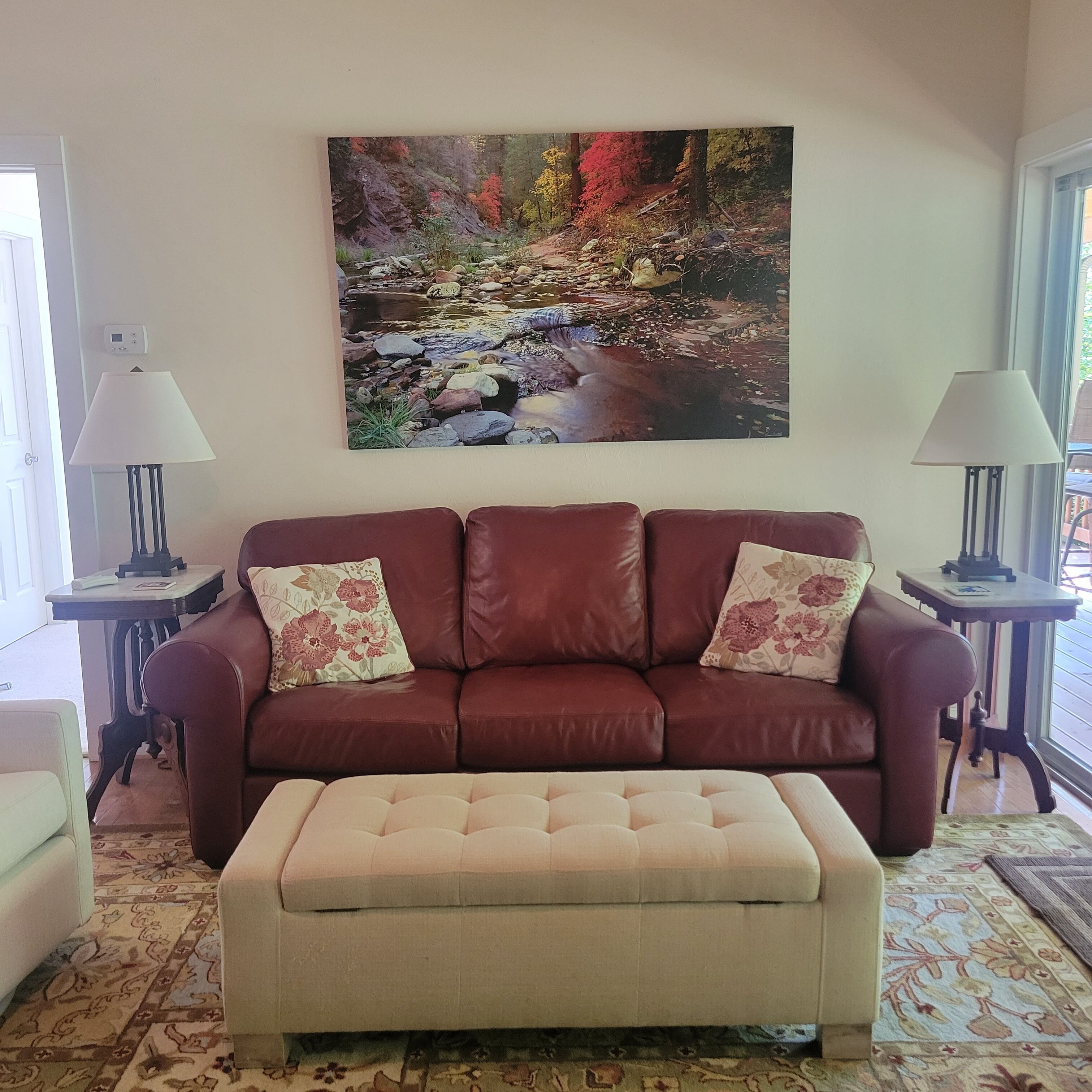 A living room with a brown leather sofa, a beige ottoman, and a large painting of a stream. There are two floor lamps on either side of the sofa.