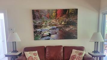 A living room with a brown leather sofa, a beige ottoman, and a large painting of a stream. There are two floor lamps on either side of the sofa.