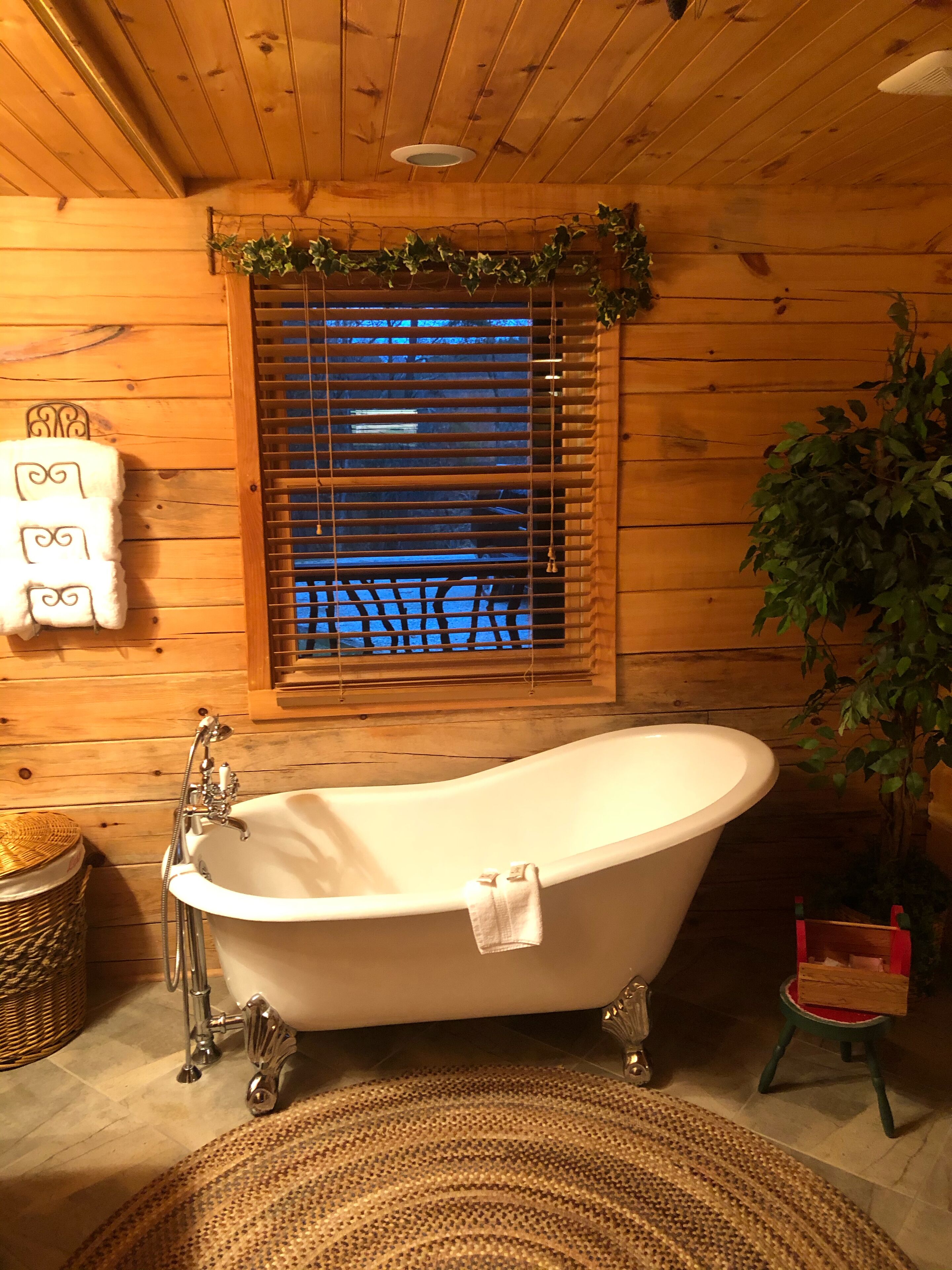 Interior de una cabaña de madera con una bañera independiente, una cesta de mimbre y una ventana con persianas.