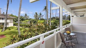 A balcony with a view of a lush garden and palm trees. There is a table with chairs and a small table with fruits.