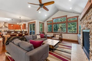A spacious living area with a large gray sectional sofa, a wooden coffee table, and a striped rug. There is a kitchen area with wooden cabinets and a stone fireplace. Large windows provide views of the surrounding trees.