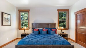 Un dormitorio con una cama grande, dos mesitas de noche, una puerta de madera y dos ventanas con vistas a árboles.