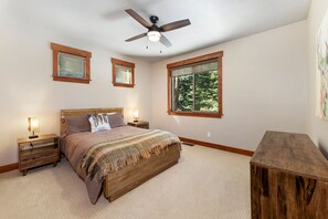 A bedroom with a wooden bed, two bedside tables with lamps, a ceiling fan, and a large window with a view of trees.