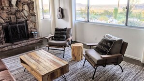 A living room with a stone wall, a fireplace, a large window with a view, and two armchairs with patterned cushions. - Stunning Views: Rockwood Hill House (Joshua Tree)