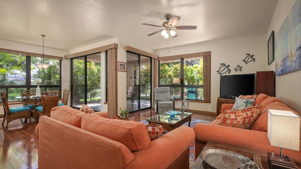 A spacious living room with a large sofa, a coffee table, and a television. There are large windows providing a view of the outdoors. -  SPECTACULAR !!  Ground Floor Condo Sleeps 5  Golf ,Tennis ,.Gym Pool, Jacuzzi (waikoloa village)