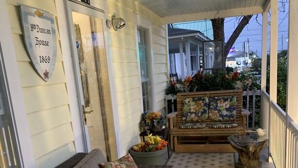 A covered porch with a beige sofa, a wooden bench with floral cushions, and a decorative birdhouse.