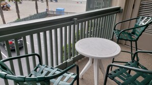 Varanda com mesa e cadeiras, com vista para uma praia com guarda-sóis e algumas pessoas.