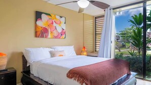 Un dormitorio con cama, ventilador de techo, mesita de noche, lámpara y ventana con vista a follaje.