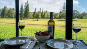Una mesa servida con una botella de vino, un cuenco de fruta y dos copas de vino, con vistas a un paisaje cubierto de hierba y árboles.