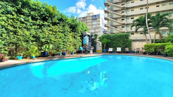 A swimming pool with lounge chairs and potted plants, surrounded by greenery and a multi-story building.