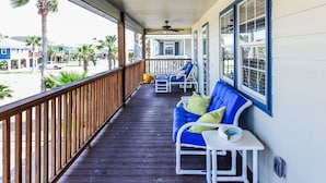 A covered porch with a wooden deck, blue cushioned seating, and a view of the beach and buildings in the background. - Twelve Palms-Pet Friendly, Golf Cart Zone, Close to Beach, Downtown, City Pool  (Port Aransas)