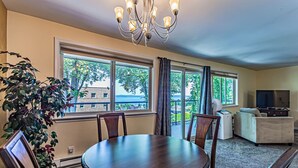 A dining area with a round table and chairs, large windows with curtains, a chandelier, a television, and a plant.