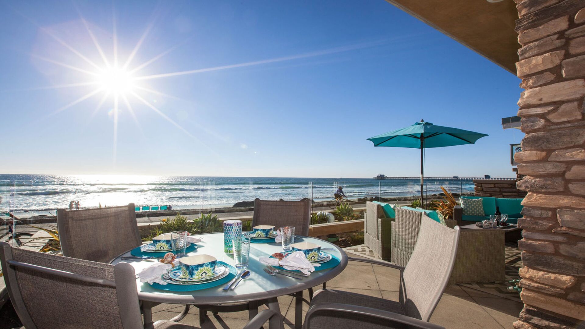 Un espace de restauration en plein air offrant une vue sur l’océan et la jetée, avec une table dressée avec de l’assiette bleue et blanche, un parasol turquoise et des chaises en osier.