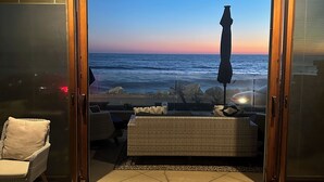 Une piĂšce avec vue sur lâocĂ©an au coucher du soleil, un canapĂ© en osier, un fauteuil et un grand parasol.