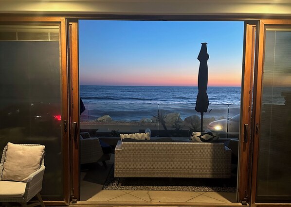 A room with a view of the ocean at sunset, a wicker sofa, a chair, and a large umbrella.