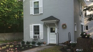 Ein zweigeschossiges Haus mit einer Haustür, zwei Fensterladen, einer kleinen Veranda und einem Garten mit Blumen und Sträuchern.