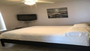 A bedroom with a bed, a ceiling fan, a flat-screen TV mounted on the wall, and a window with blinds.