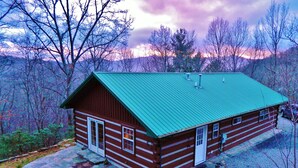 Une cabane en rondins surmontée d’un toit végétalisé, avec une entrée pavée de pierres et une allée gravillonnée.
