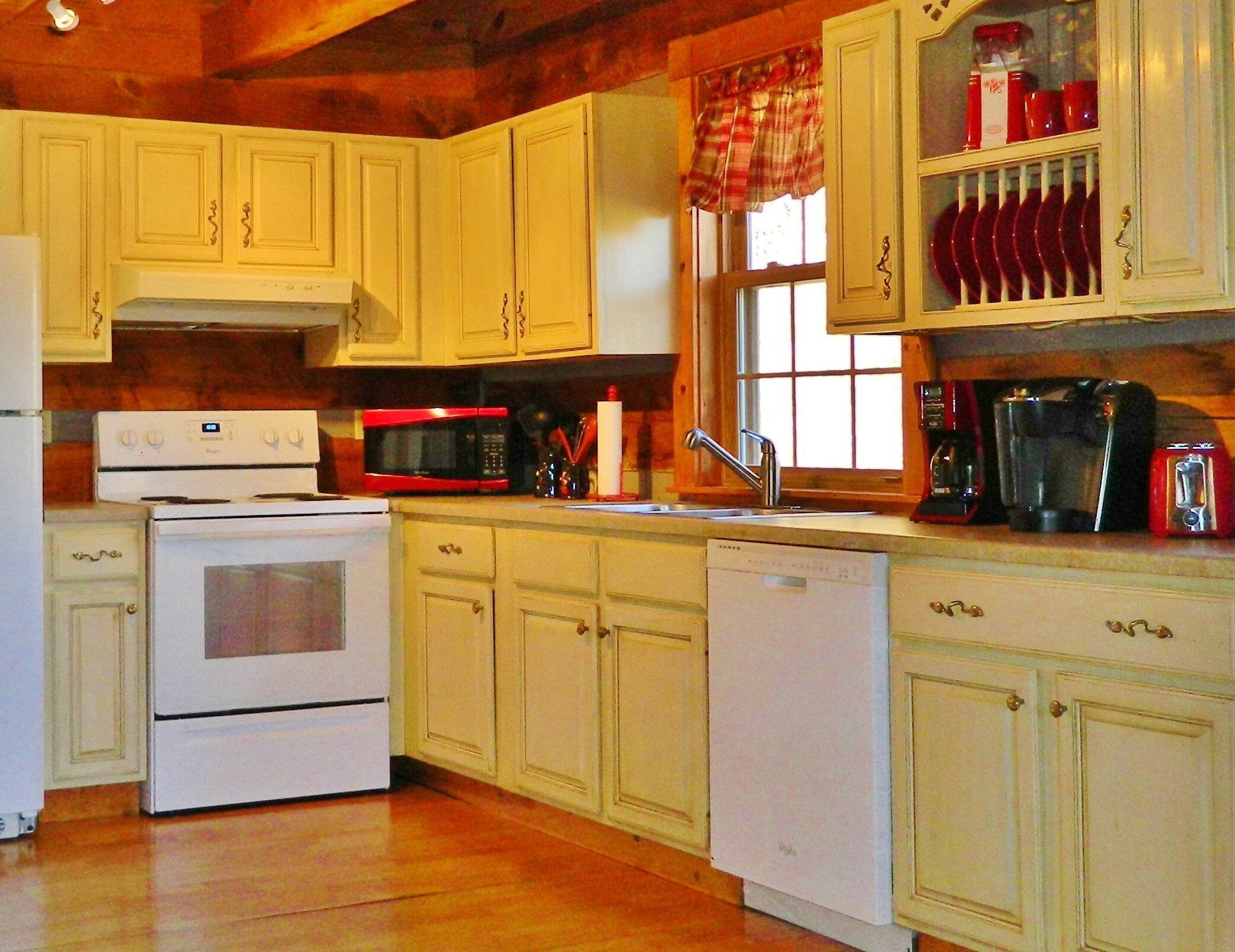 A kitchen with white cabinets, a red microwave, a stove, a dishwasher, and a window with plaid curtains.